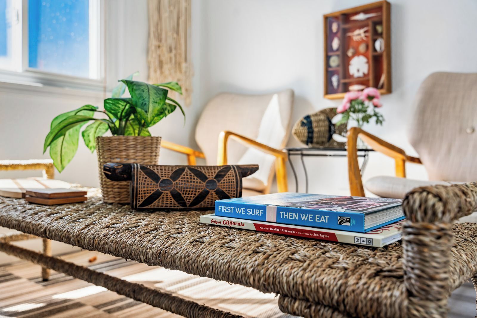 A wicker sofa with a woven pattern, a potted plant, and two books titled 'FIRST WE SURF' and 'THEN WE EAT' on the sofa.