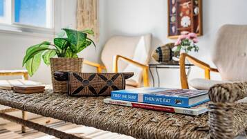 A wicker sofa with a woven pattern, a potted plant, and two books titled 'FIRST WE SURF' and 'THEN WE EAT' on the sofa.