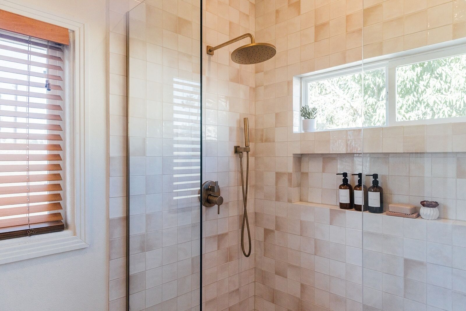 A glass-enclosed shower with a handheld showerhead, a window with blinds, and a shelf with toiletries.