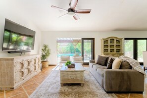 Un salón con televisión de pantalla plana, ventilador de techo, sofá, mesa de centro y vistas a una piscina.