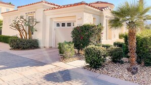 A two-story house with a tiled roof, a garage door, and a well-maintained garden with various shrubs and a palm tree.