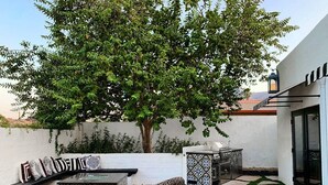 An outdoor patio with a seating area, a grill, and a tree providing shade.