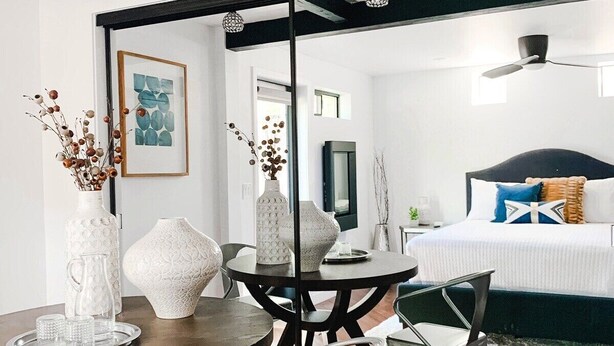 A modern interior with a dining area featuring a round table and chairs, a bed with a dark headboard, and decorative vases.