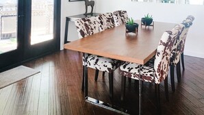 A dining area with a wooden table and cow-print upholstered chairs, a black-framed picture on the wall, and large windows with a view of greenery.