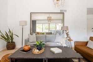 Uma sala de estar moderna com um sofá cinzento, uma mesa de centro com objetos decorativos, um espelho grande e um candelabro.