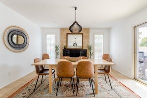 Uma sala de jantar com uma mesa de madeira, quatro cadeiras castanhas, um tapete estampado, uma luminária pendente e uma lareira.