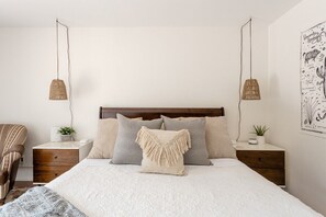Quarto bem arrumado com uma cama grande, duas mesas de cabeceira, uma cadeira e uma obra de arte na parede.