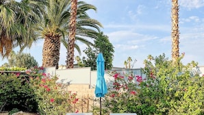 Una veranda con due poltrone bianche imbottite e un ombrellone blu, circondata da palme e cespugli fioriti.