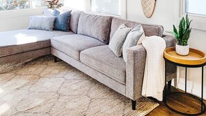 Een moderne woonkamer met een grijze modulaire bank, een rond houten bijzettafeltje, een plafondventilator en een groot raam met bamboeblamen.