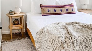Een slaapkamer met een bed, nachtkastjes, een raam met een bamboe zonwering, en diverse geweven manden en tassen aan de muur.