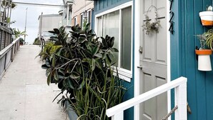 Een smalle straat met huizen, waarvan er één een witte deur en teal gevelbekleding heeft, en een plantenbak met groene planten ervoor.