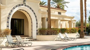 A poolside area with lounge chairs, a table, and a building with an arched entrance. - Casita Azul - Resort Amenities, Tennis 2BR260196 (La Quinta)
