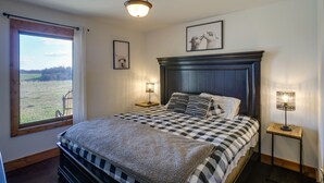 A bedroom with a large bed, two bedside lamps, a window with a view of a field, and a framed picture on the wall. - Rustic Missouri Vacation Rental w/ Hill Views! (Greenfield)