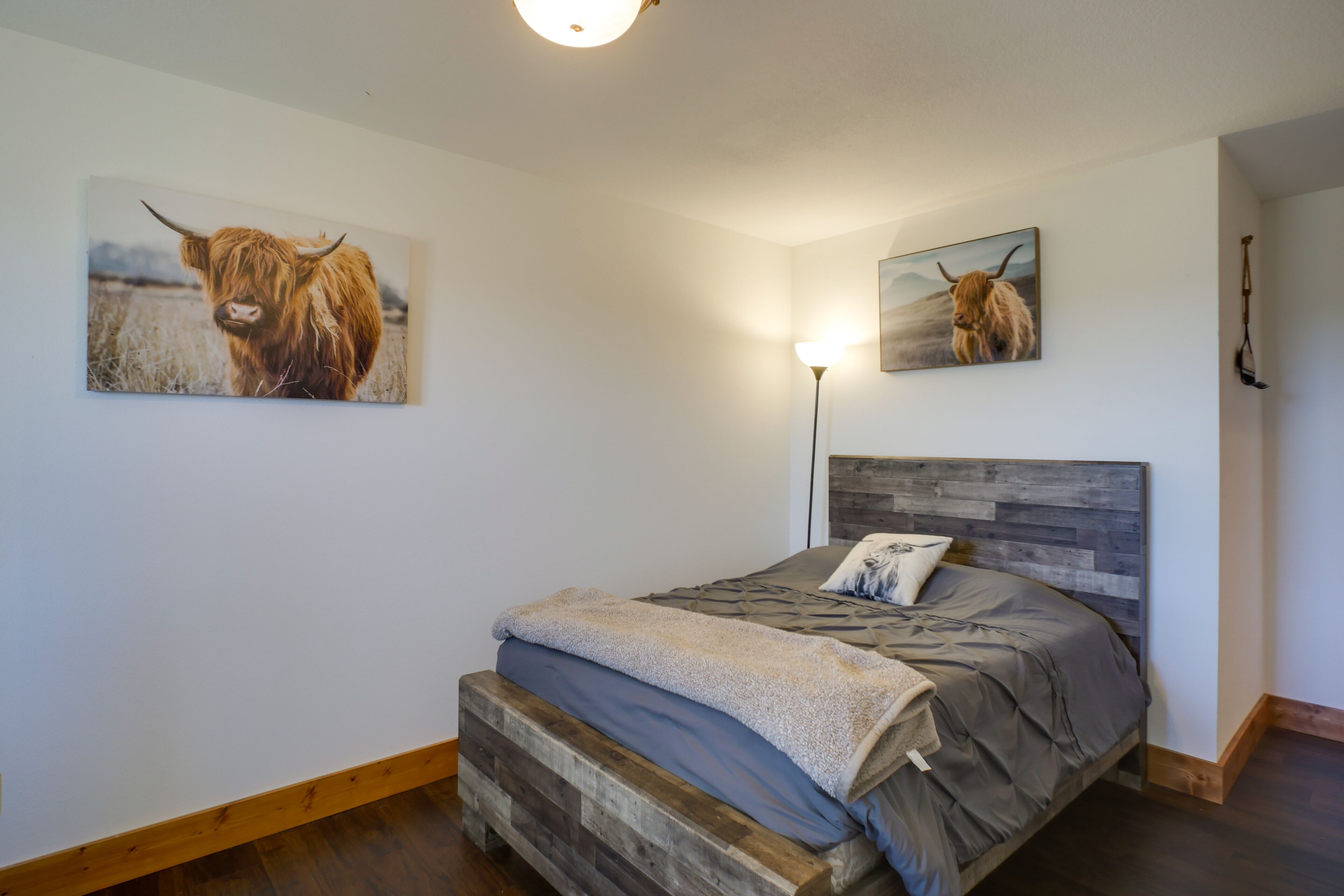 A bedroom with a wooden headboard, a bed with grey bedding, and two paintings of a Highland cow on the walls.
