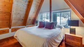 A bedroom with a sloped ceiling, a large bed, a nightstand, and a window with a view of trees. - Donner Lake Views: Rustic Truckee Cabin! (Truckee)