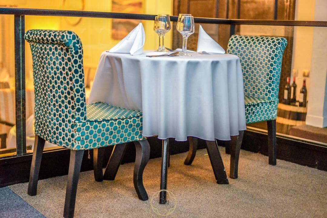A dining area with two patterned chairs and a round table, each with a white tablecloth and a pair of wine glasses.
