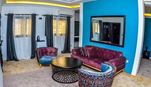 A modern living room with a purple sofa, two armchairs, a coffee table, and a patterned ottoman. - AMJ Hotel (Yaoundé)