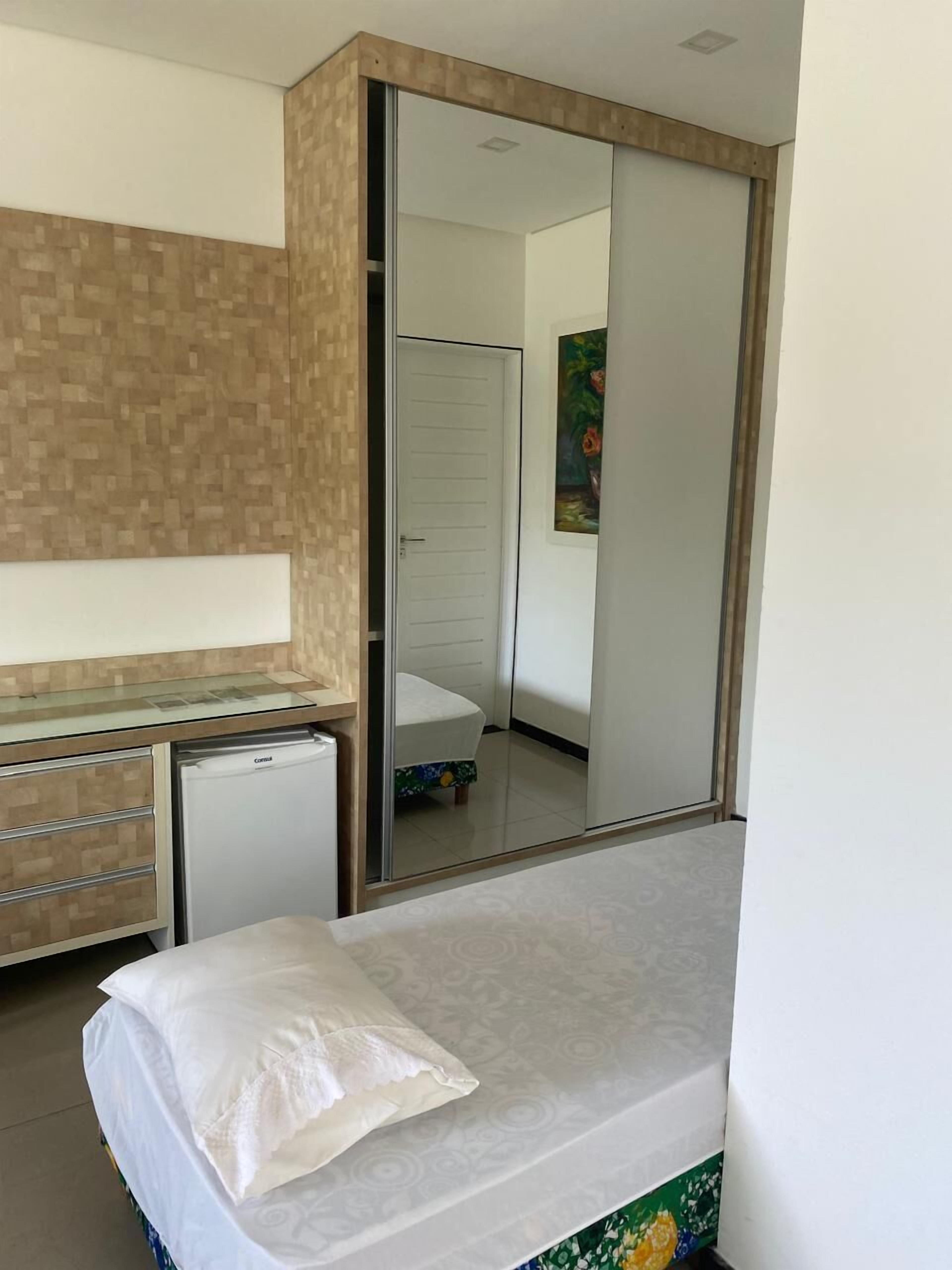 Quarto com cama, pequena mesa com frigorífico, guarda-roupa e espelho.