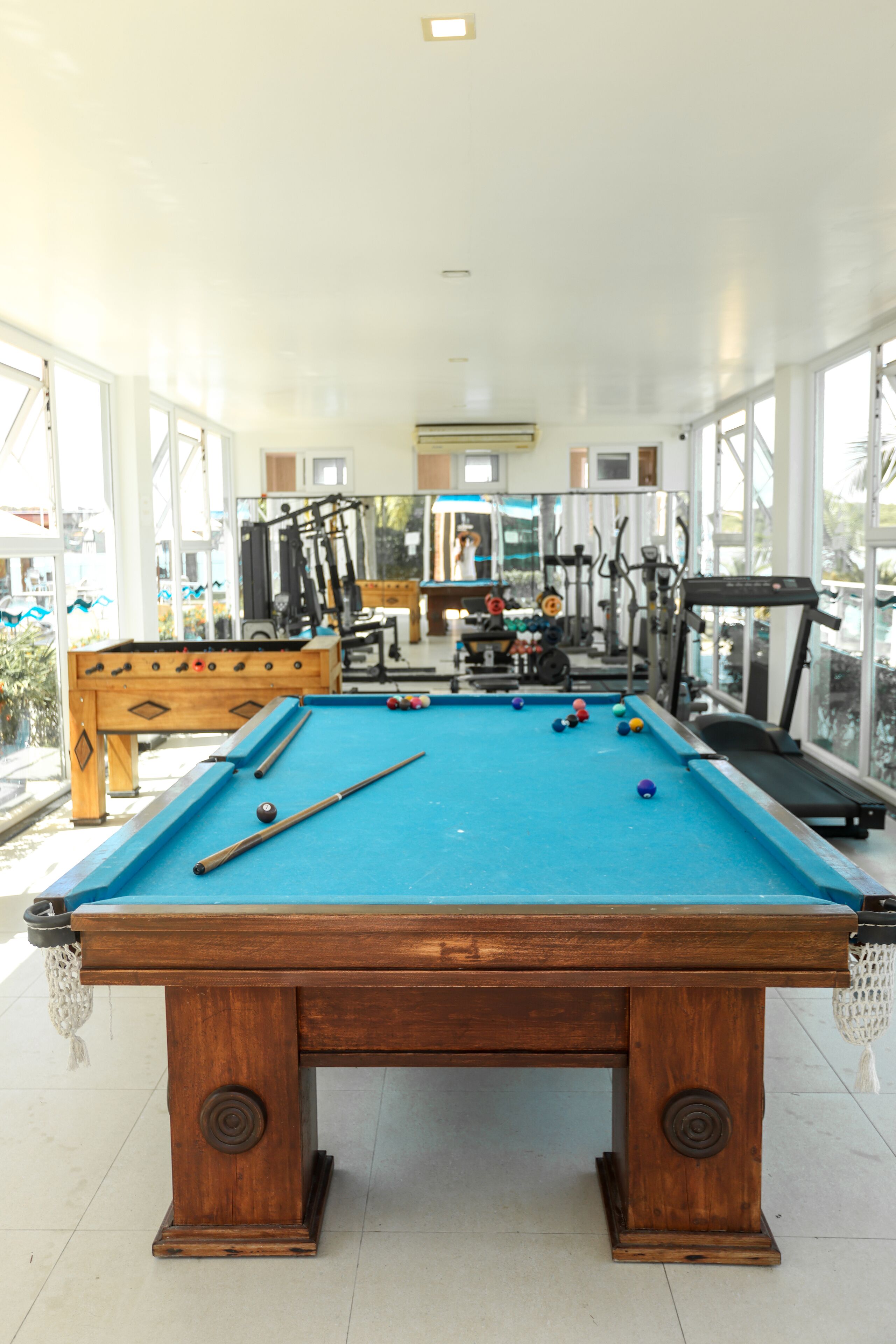 A pool table with billiard balls, a foosball table, and exercise equipment in a bright room with large windows.