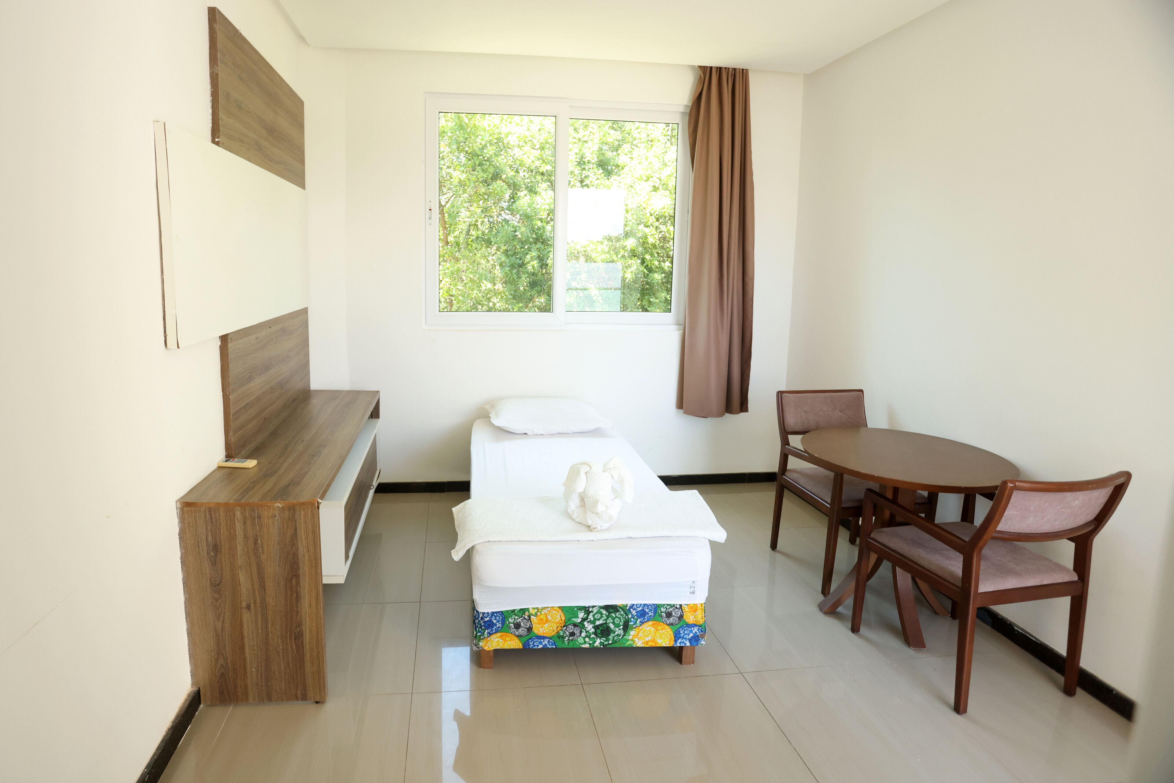 Een kleine, schone kamer met een bed, een houten bankje, een eettafel met stoelen en een raam met gordijnen.