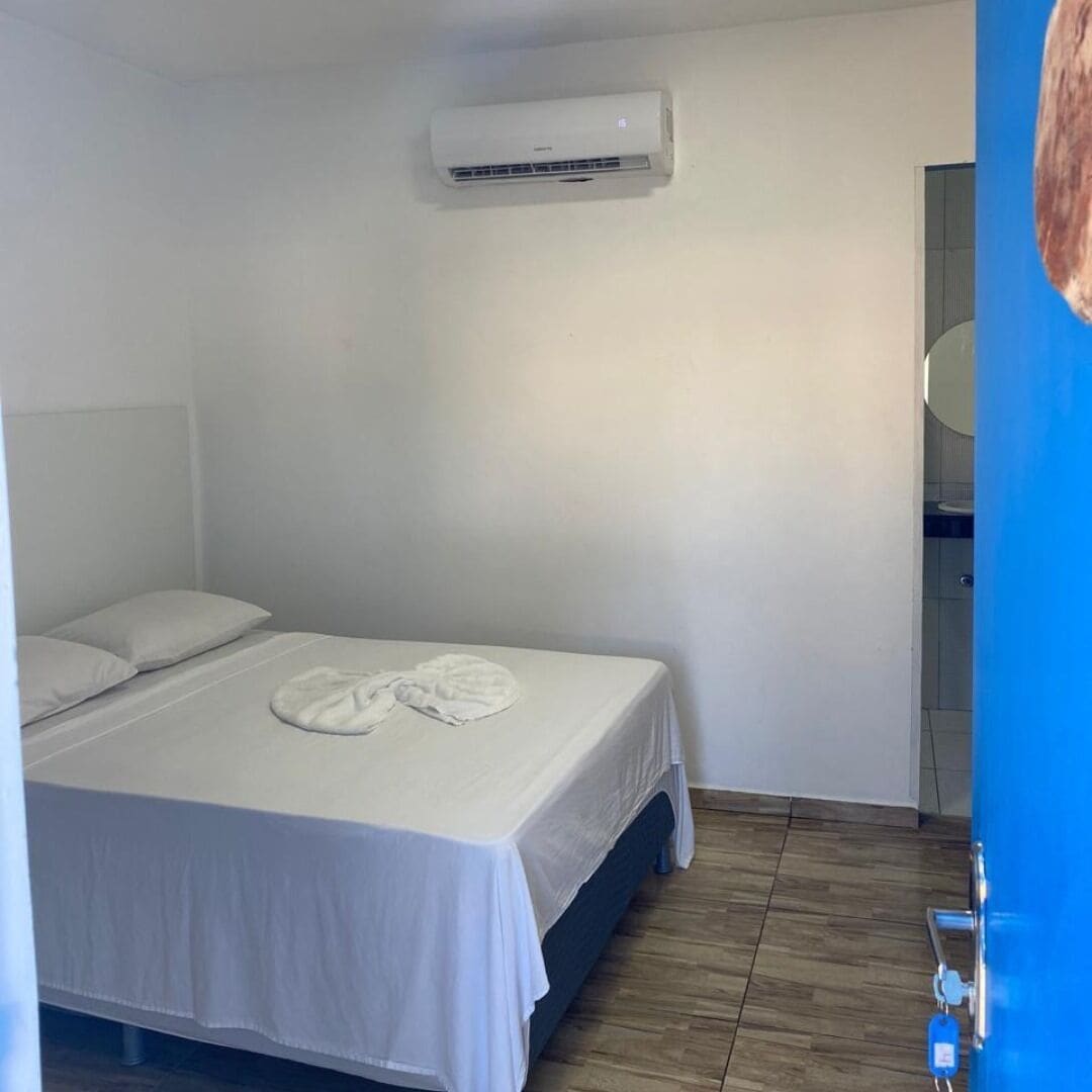 A hotel room with a bed, white linens, a wall-mounted air conditioner, and a wooden floor.