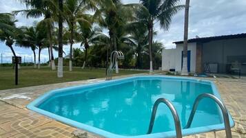 Piscina com deck ladrilhado, cercada por palmeiras e um edifício com telhado em azulejos.