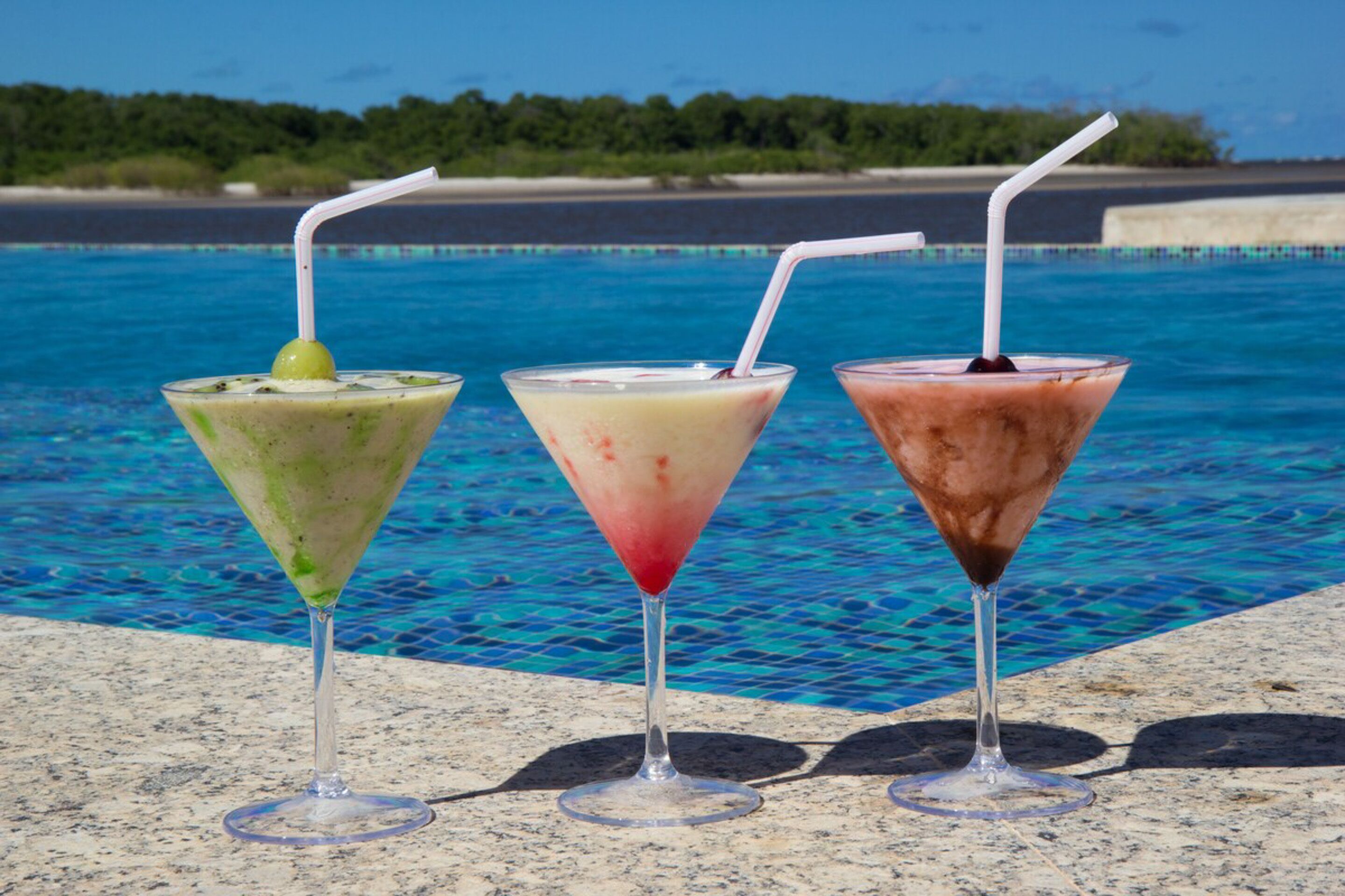 Three cocktails in martini glasses with straws, placed on a stone surface by a pool with clear blue water and a view of the beach and trees in the background.