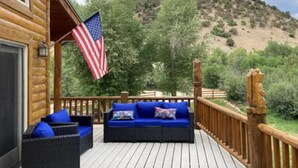 Una terraza de madera con un sofá y cojines, una bandera estadounidense y vistas a un paisaje montañoso.