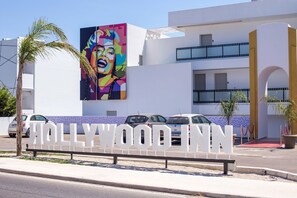 Entrada de un hotel con un gran letrero que dice 'Hollywood Inn', fachada de edificio moderno y un área de estacionamiento con coches.