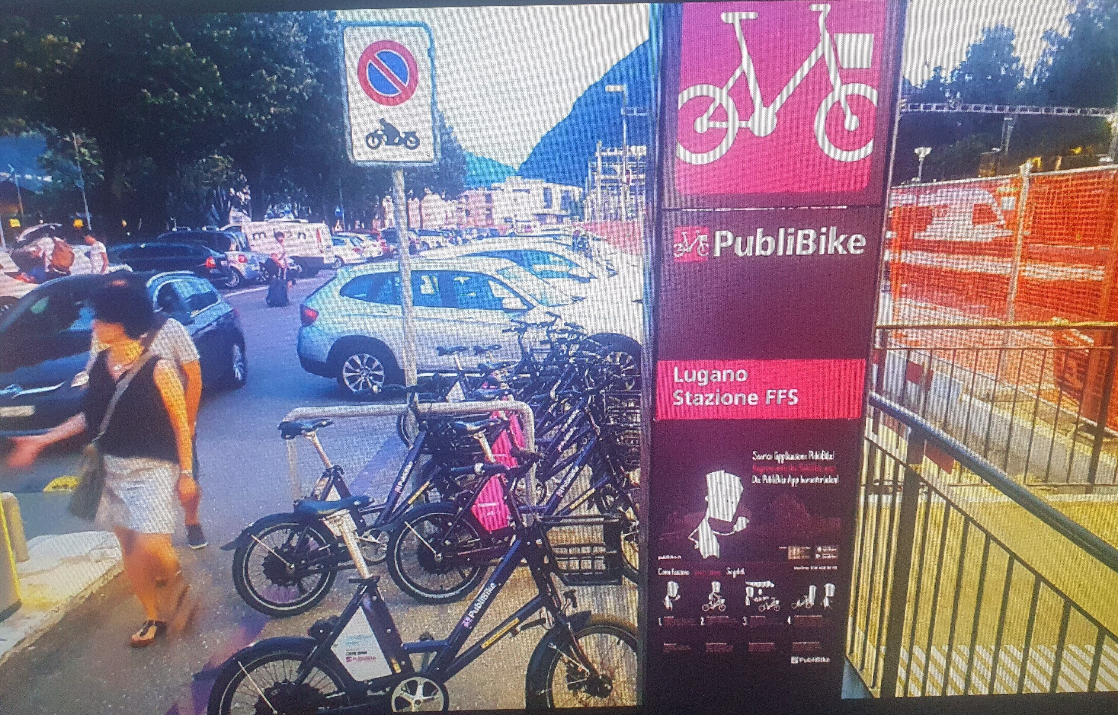 Een drukke straat met geparkeerde fietsen, een parkeerverbod en een reclame voor een fietsenstalling.