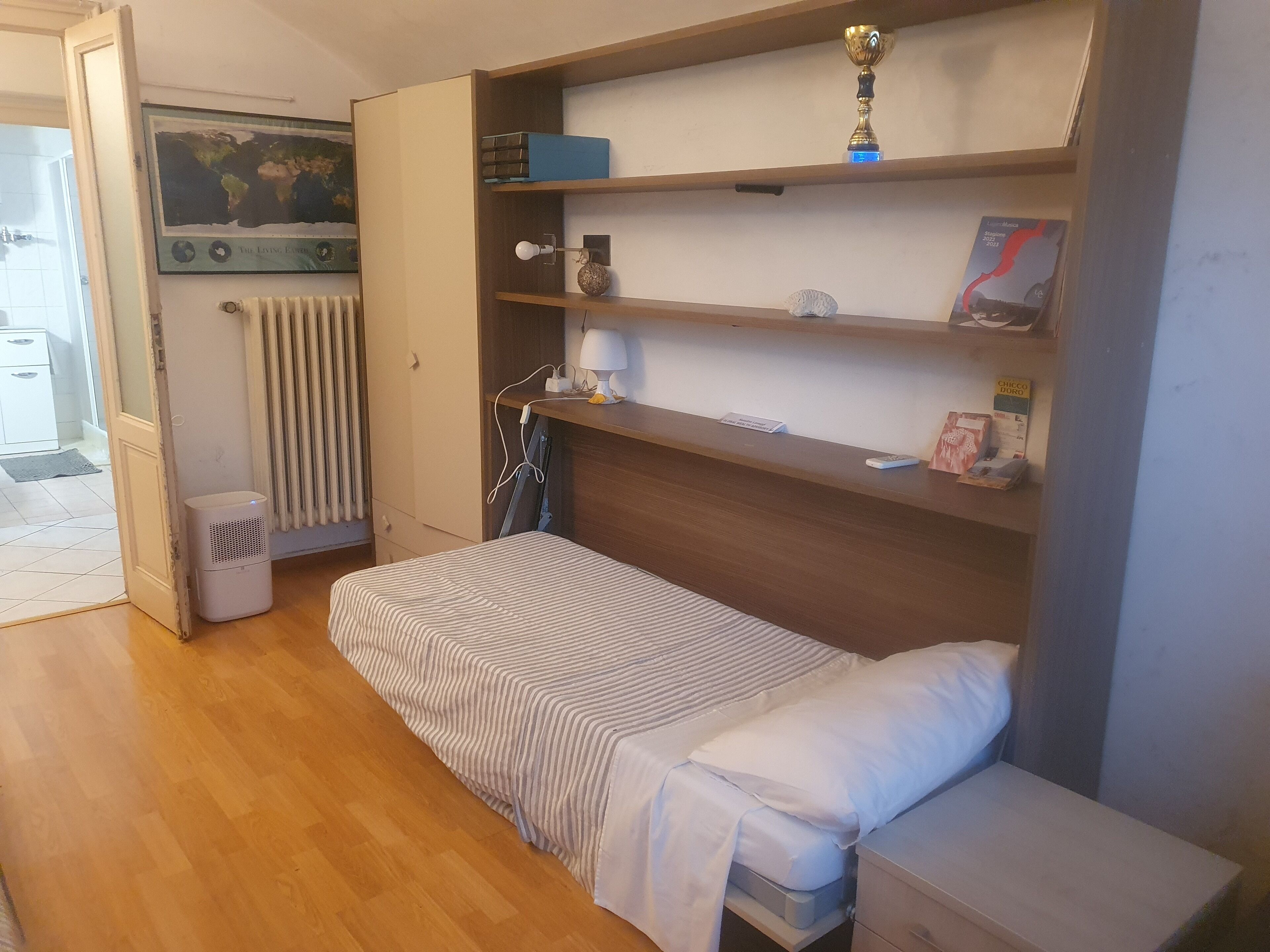 A compact bedroom with a bed, a small bedside table, a shelf with a trophy, and a radiator.