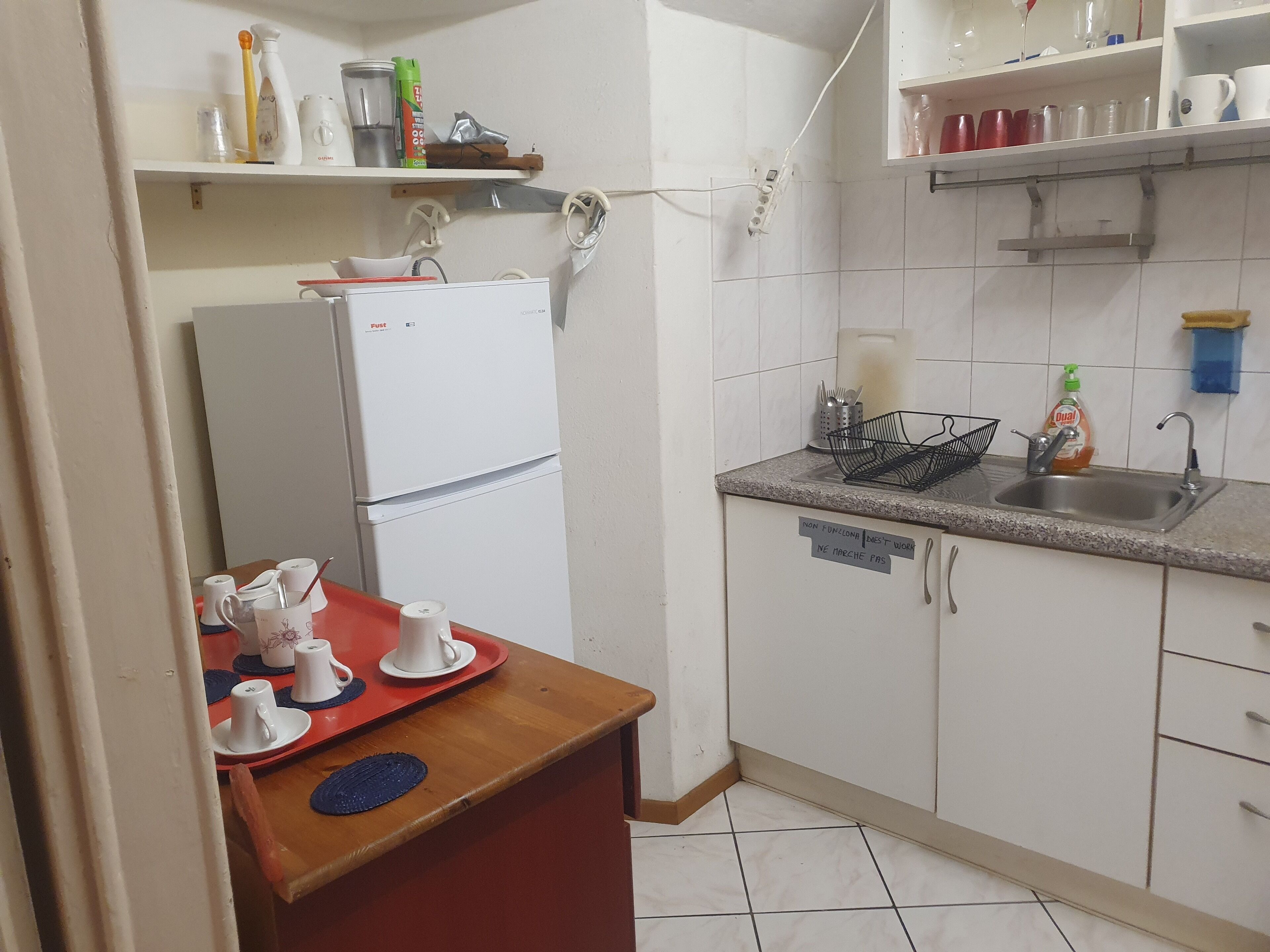 Une cuisine compacte équipée d’un réfrigérateur, d’un évier et de divers ustensiles de cuisine rangés sur des étagères et sur le plan de travail.