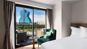 A hotel room with a large bed, a green armchair, a small round table, and a view of a building through a large window.