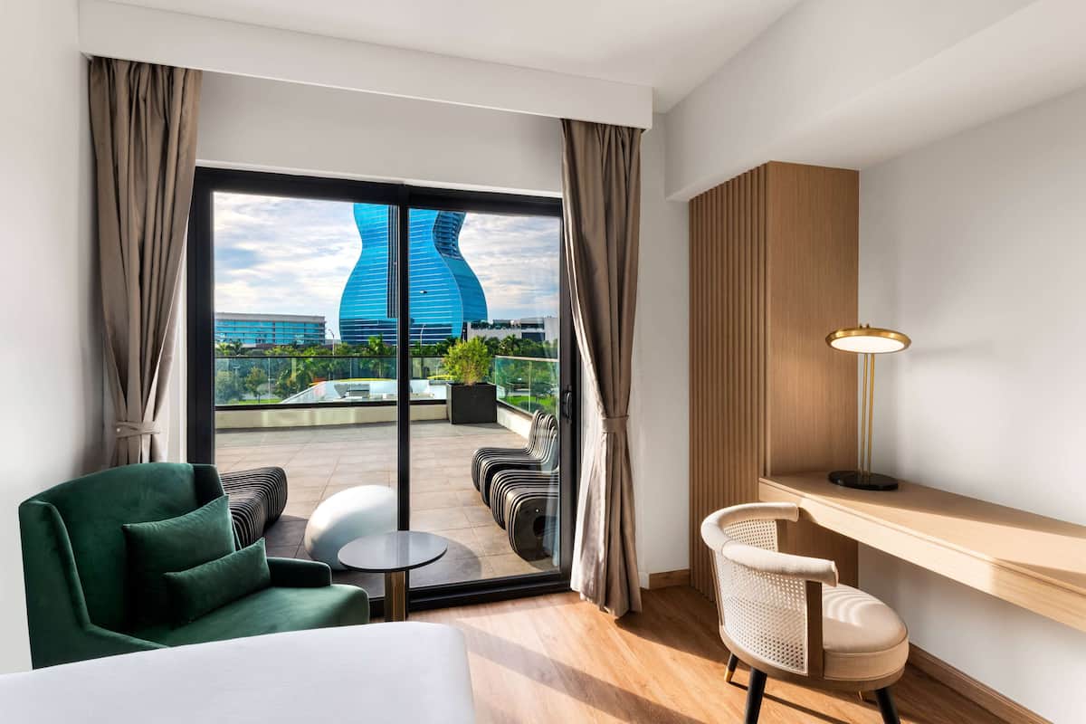 Habitación de hotel moderna con un amplio ventanal corredero que da a un balcón, un sillón verde, una pequeña mesa redonda, un escritorio con lámpara y vistas a un edificio singular.