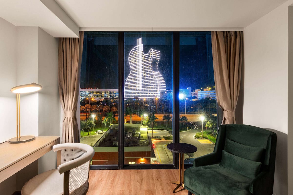 Una habitación de hotel moderna con un ventanal grande, un escritorio, una silla y un sillón verde.