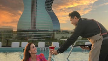 Una persona entrega una bebida a otra junto a una piscina, con un edificio con forma de guitarra al fondo.