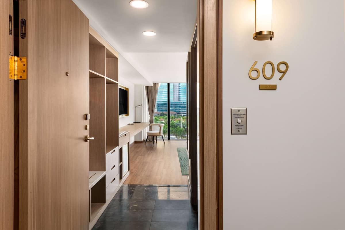 Una habitación de hotel moderna con un armario de madera, un escritorio, una silla y vistas a la ciudad a través de amplios ventanales.