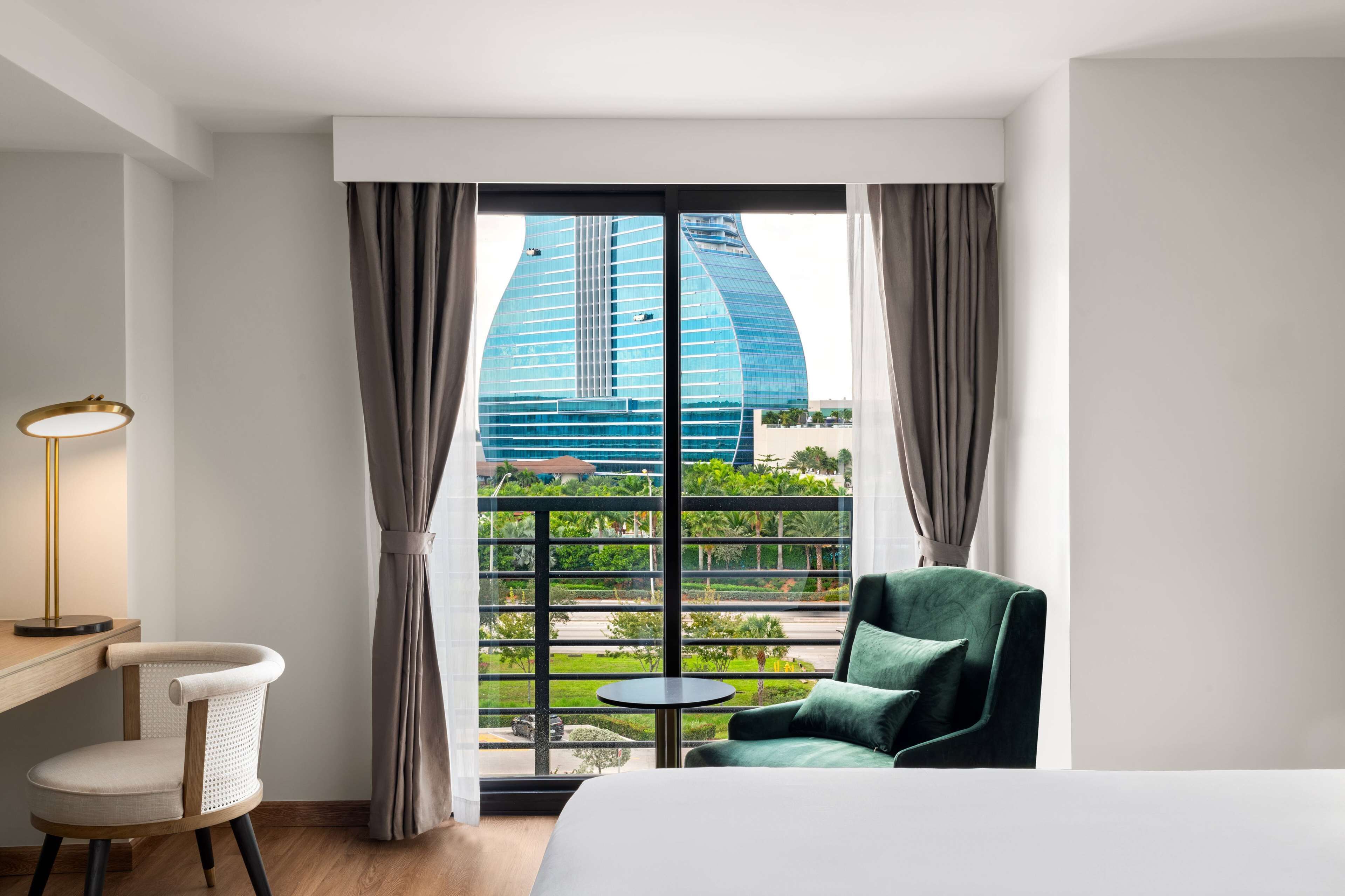 A hotel room with a large window, a bed, a chair, a small table, and a lamp.