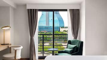 A hotel room with a large window, a bed, a chair, a small table, and a lamp.