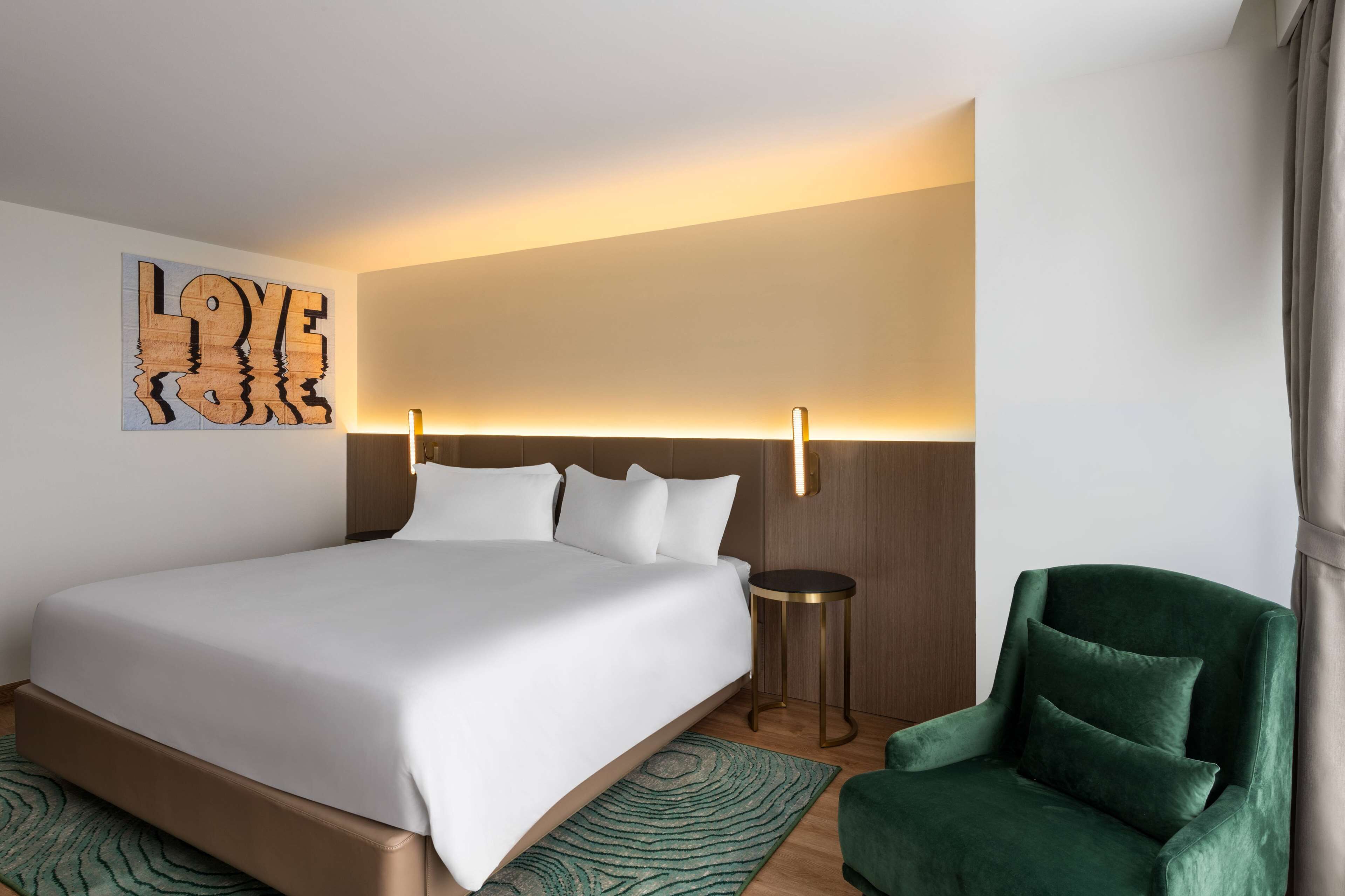 A modern hotel room with a large bed, a green armchair, a round side table, and a wall-mounted artwork with the word "LOVE".