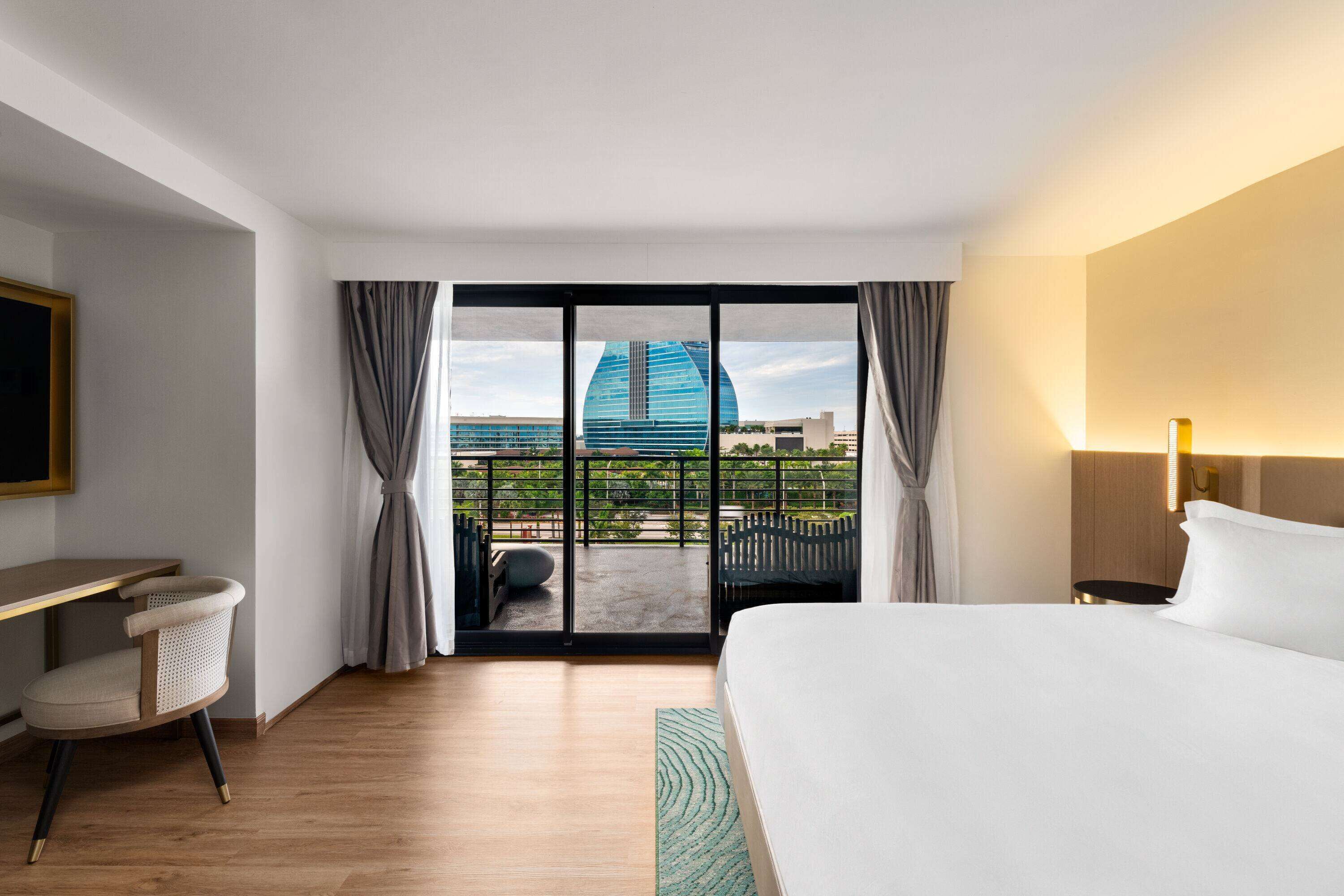 A hotel room with a large bed, a desk, and a view of a modern building through the glass door.
