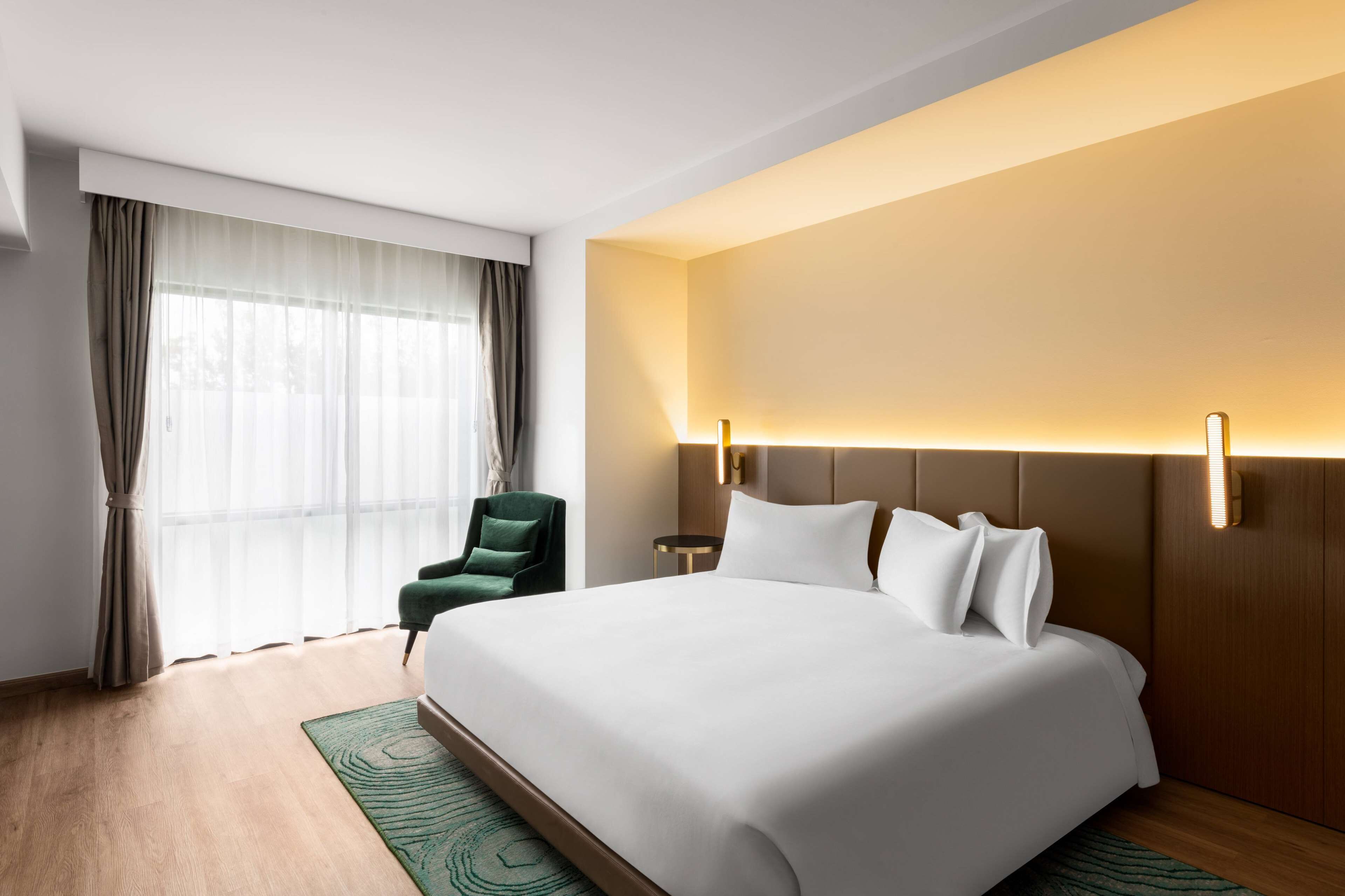 A hotel room with a large bed, a green armchair, a nightstand, and a wall-mounted lamp.