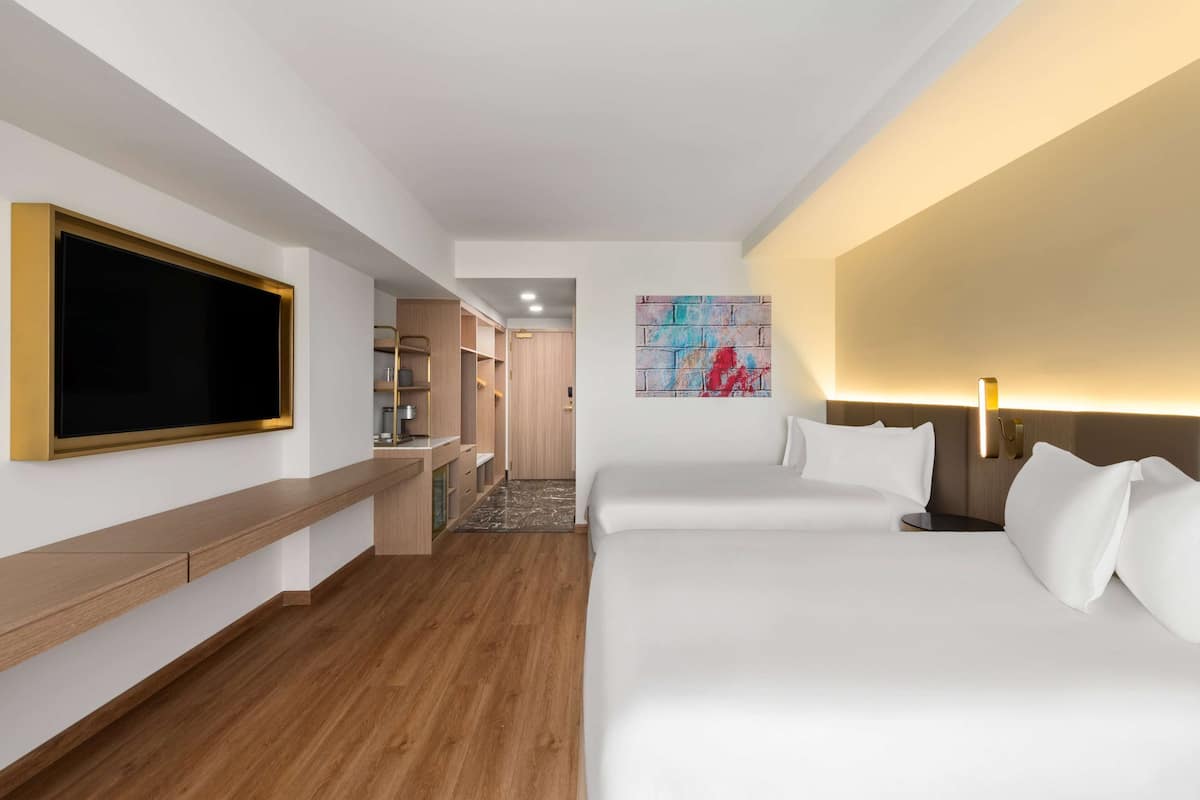 Habitación de hotel con un televisor de pantalla plana grande, una cama, una mesita de noche, un cabecero con iluminación integrada y una obra de arte abstracta y colorida en la pared.