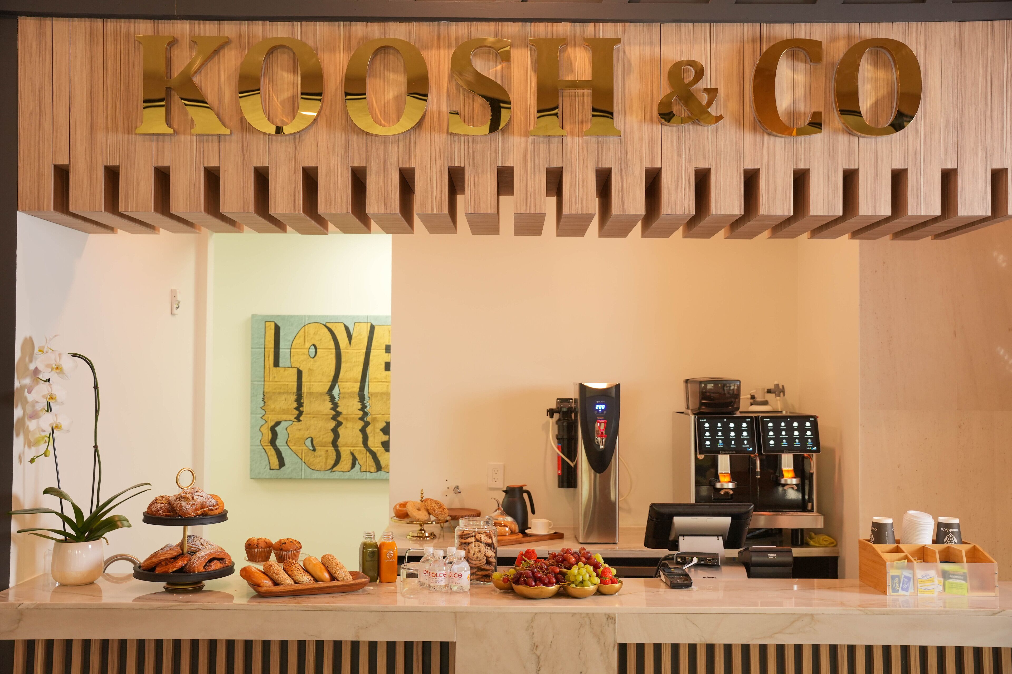 A coffee shop counter with a sign reading "KOOSH & CO", a coffee machine, pastries, and a fruit bowl.
