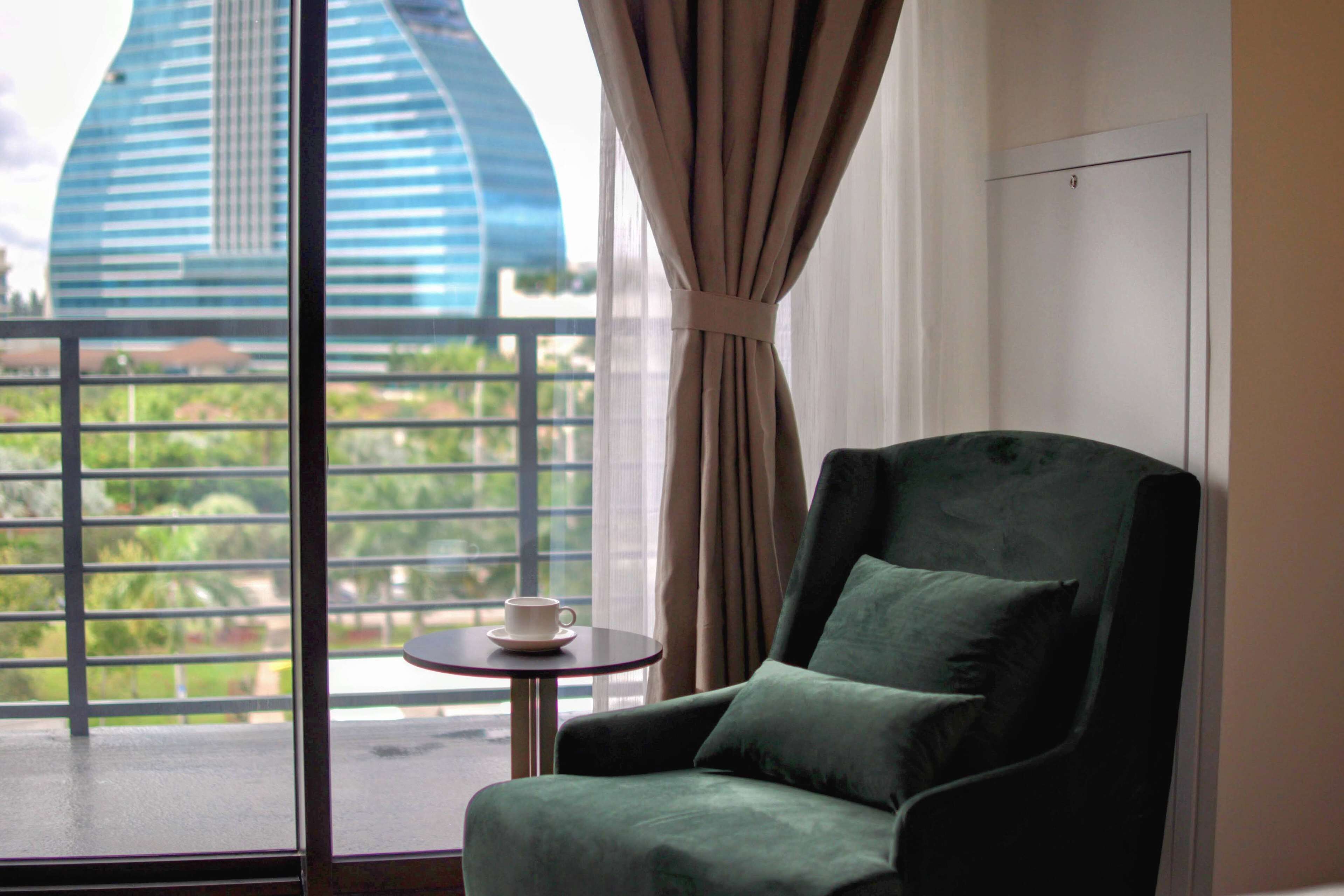 A room with a view of a modern building, a green armchair, and a small table with a cup.