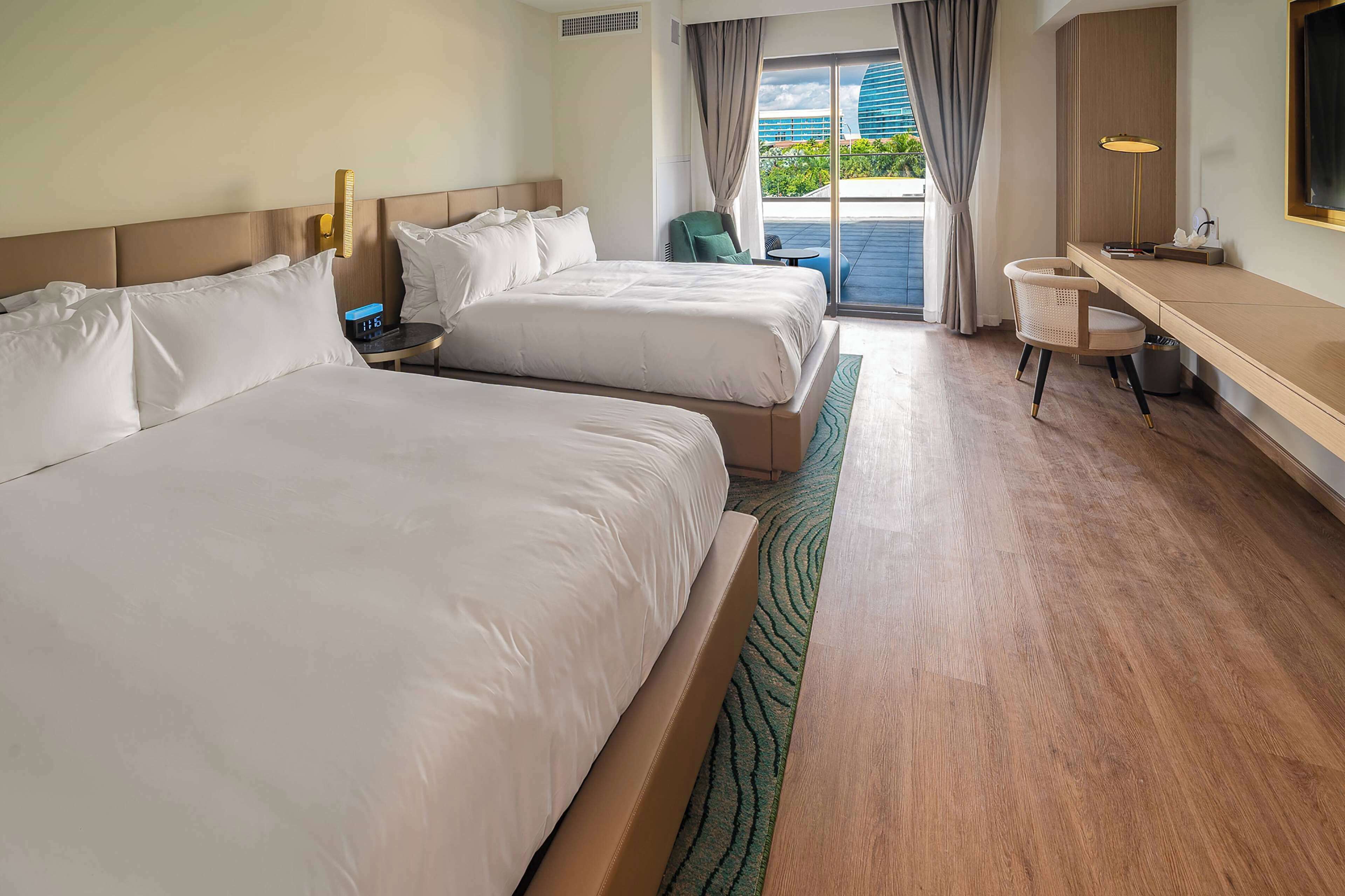 A hotel room with two beds, a desk, a chair, and a view of the outdoors.