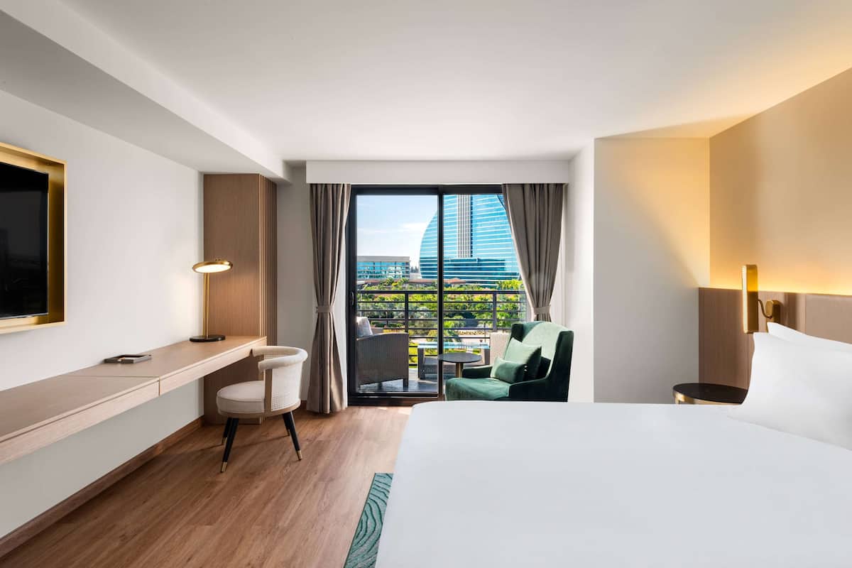 Habitación de hotel con cama, escritorio, silla, televisor, lámpara y balcón con vistas a un edificio.