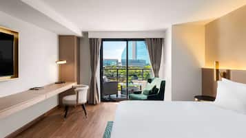 Habitación de hotel con cama, escritorio, silla, televisor, lámpara y balcón con vistas a un edificio.