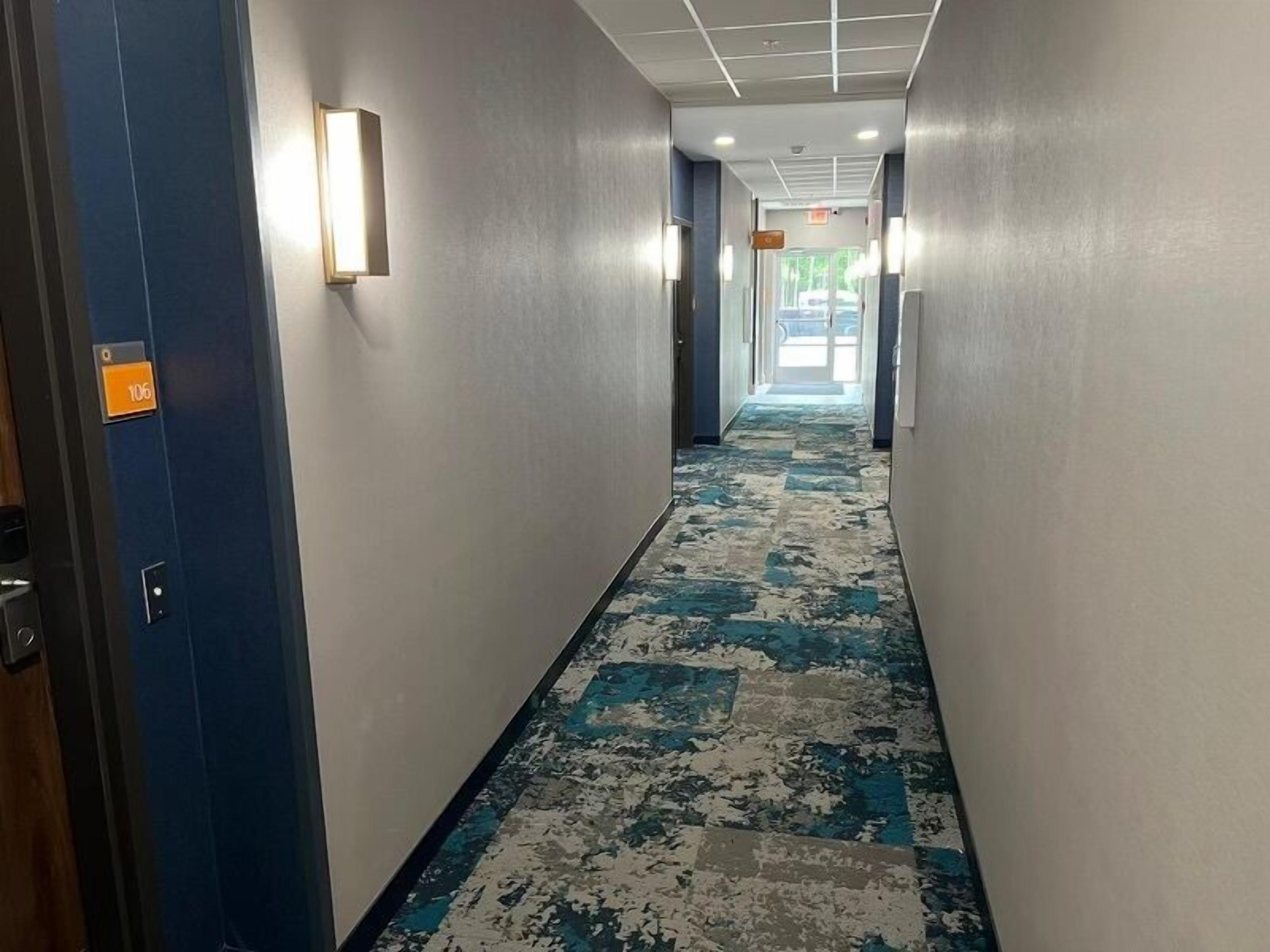 A hotel hallway with a patterned carpet, light fixtures on the walls, and a door with a number sign.