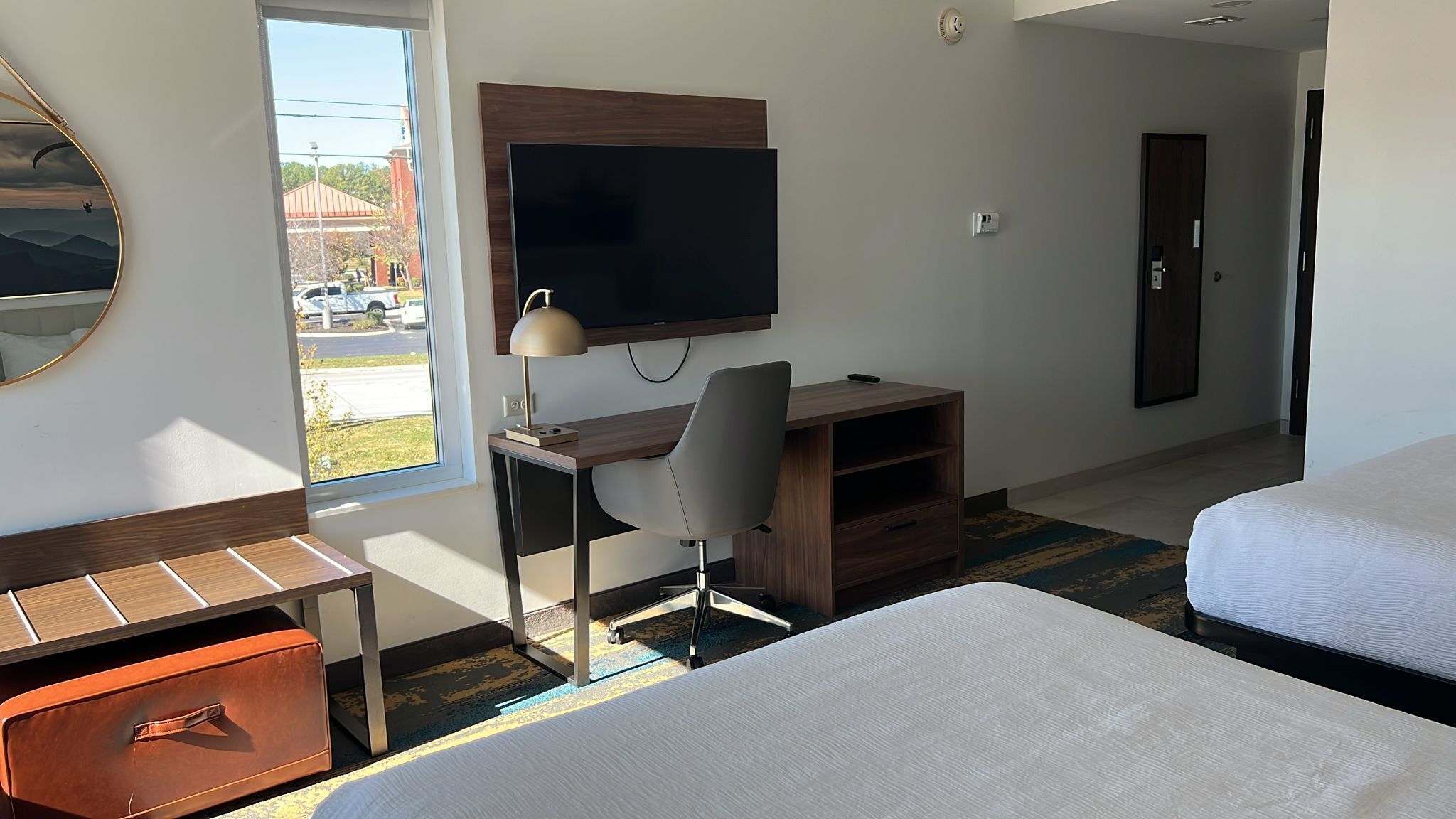 Hotelkamer met een bed, een bureau met stoel, een televisie en een raam met uitzicht op een parkeerplaats.